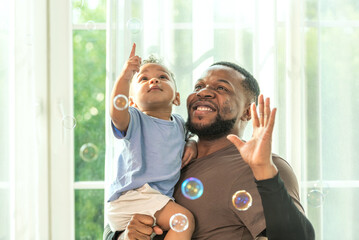 Happy african black parents dad father throw baby son on piggyback and neck riding teasing fun near window. Black baby son and daddy enjoy teasing kiss cheek and neck riding at window light