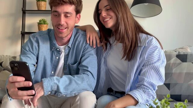 Happy couple sitting together on sofa at home, using smartphone and smiling while spending time together, concept of love, relaxation, communication, modern lifestyle and positive relationship