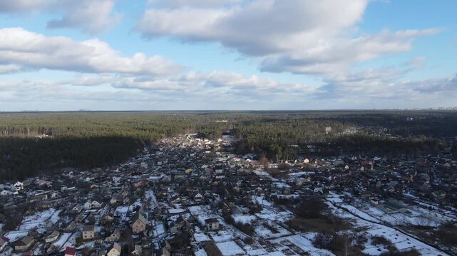 Aerial panoramic view of a small town partly covered in snow, located next to a dense forest. Shot on a sunny winter day with blue sky and scattered clouds. High-altitude drone footage.