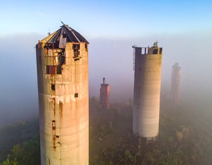 Foggy morning landscape showing abandoned industrial silos and a crumbling building under a moody sky.