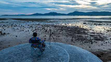 A man sits alone eating in a folding chair on a stone platform, overlooking the rocky intertidal flats, camera at his side, and the peaceful sea in the distance. A quiet moment of solitude at sunset.
