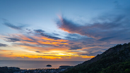 A breathtaking sunset paints the sky in vivid hues of orange and blue over a coastal town, with lush green hills framing the scene. A distant hilltop structure adds a serene tranquil landscape.