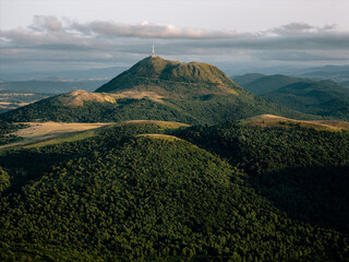 Expansive aerial shot of Puy de D&ocirc;me volcano surrounded by rolling green hills, dramatic clouds, and soft golden sunlight, capturing the tranquil mood of Auvergne&rsquo;s volcanic landscape.
