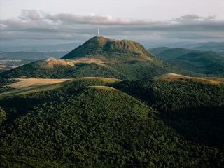 Expansive aerial shot of Puy de Dôme volcano surrounded by rolling green hills, dramatic clouds, and soft golden sunlight, capturing the tranquil mood of Auvergne’s volcanic landscape. © Pavlo Glazkov