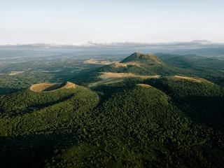 Aerial photo of Chaîne des Puys in Auvergne, France, showing volcanic craters, the Puy de Dôme summit, dense forests, and soft sunset light over a tranquil landscape. © Pavlo Glazkov