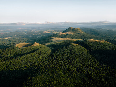 Aerial photo of Cha&icirc;ne des Puys in Auvergne, France, showing volcanic craters, the Puy de D&ocirc;me summit, dense forests, and soft sunset light over a tranquil landscape.