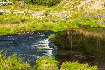 Scenic river landscape with lush green banks, gentle rapids, and a two figure walking by the water under a clear sky