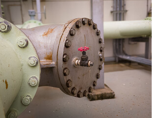 Close-up of a large industrial water pipe with bolted flange and red-handled valve. Visible rust and wear suggest active use in a mechanical or utility room.