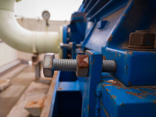 Close-up of a large bolt with dual nuts securing a motor base in an industrial setting. Rust and wear indicate active use in a mechanical system.