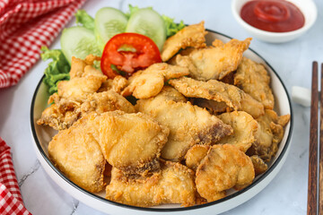 Golden-brown crispy fried fish pieces, served in a white plate with lettuce and slices of tomato and cucumber. A small red bowl of sauce is visible in the background.