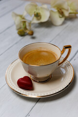 A cup of espresso on a light wooden table with a candy on a plate and an orchid flower in the background. 
