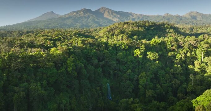 Lombok, Indonesia: Lush green rainforest covers rolling hills with serene waterfall cascading down a verdant cliffside, majestic Mount Rinjani visible in the distant background. Aerial drone panorama