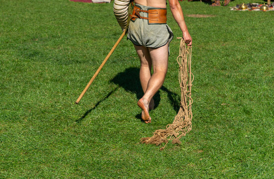 Historical reenactor in period short costume carrying net and wooden pole during outdoor event, rear view on green grass under bright sunlight.