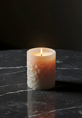 A decorative, beige candle with textured design sits on a dark gray marble surface. Burning candle with intricate design against a dark background.