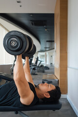 Asian male lifting dumbbells vertically on bench in gym