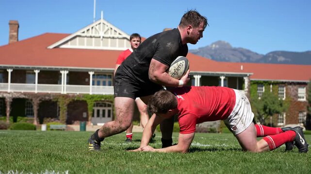 Dynamic low-angle wide video captures young adult male rugby players in black/red, intensely tackling on sun-drenched grass field, university/mountains in background, adrenaline-fueled sports