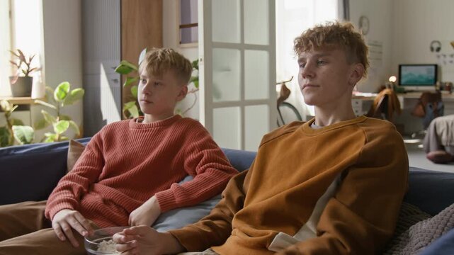 Tilt up shot of two teenage boys watching TV together and switching channels fighting for TV remote on couch