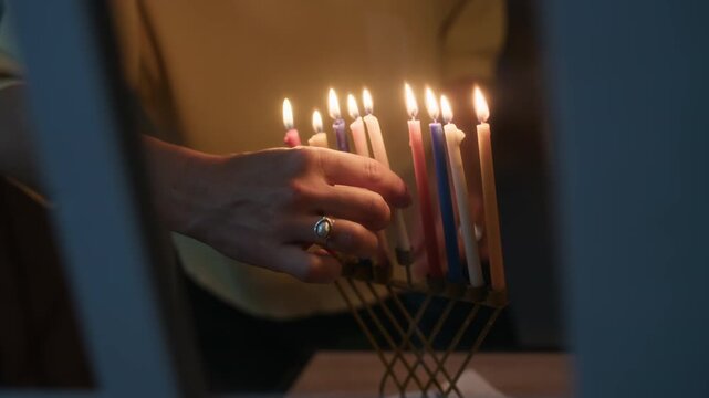Cropped close up shot of hands of unrecognizable woman placing shammash on hanukkiah with nine burning candles, celebrating final day of Hanukkah at home