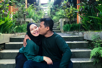 Fototapeta premium The couple sat on the steps of a lush garden; the man hugged and kissed the cheek of the woman wearing a headscarf, who smiled broadly.