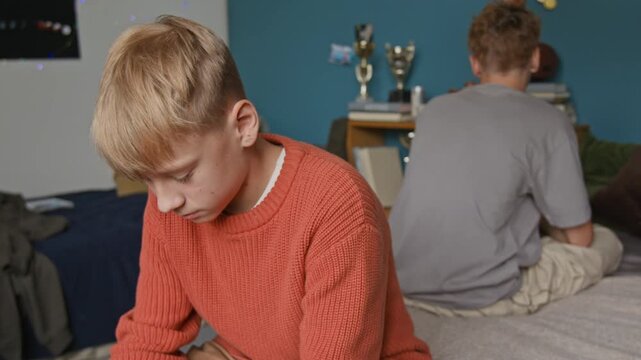Tilt up shot of two teenage boys as brothers fighting while sitting on bed at home with backs to each other