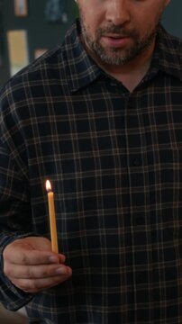 Vertical tilt down shot of young Jewish man holding burning shammash and lighting up candles on hanukkiah while singing holiday hymn Maoz Tzur, celebrating Hanukkah at home