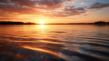 A serene sunset over a calm water body, with vibrant orange and yellow hues reflecting on the surface, framed by silhouetted trees and a clear sky.