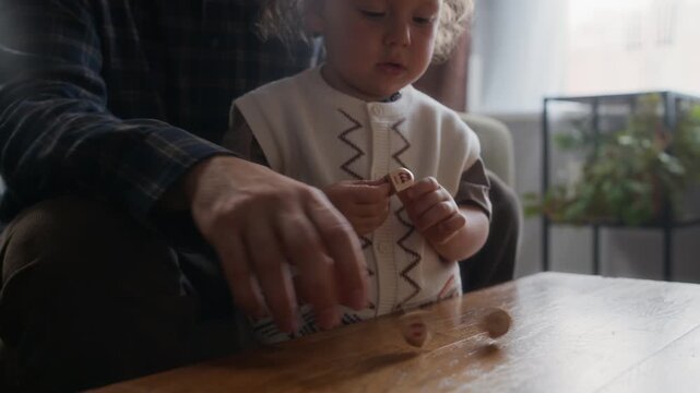 Cropped slowmo shot of hands of unrecognizable Jewish father sitting at table with his toddler son and spinning dreidels at Hanukkah