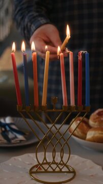 Vertical close up slowmo of hands of unrecognizable man holding burning shammash lighting up candles on hanukkiah, celebrating Hanukkah at home