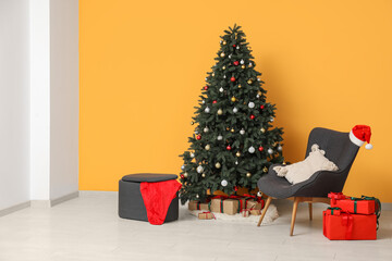 Decorated Christmas tree with gift boxes, armchair, Santa hat and pouf near yellow wall