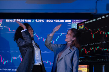 Two excited female traders celebrating a successful investment, raising arms in victory in front of stock charts.