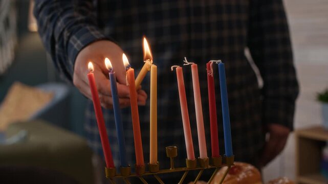 Cropped close up slowmo of hands of unrecognizable man holding burning shammash lighting up colored candles on hanukkiah, celebrating Hanukkah at home