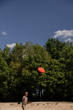 Man throws orange frisbee in park with trees and blue sky