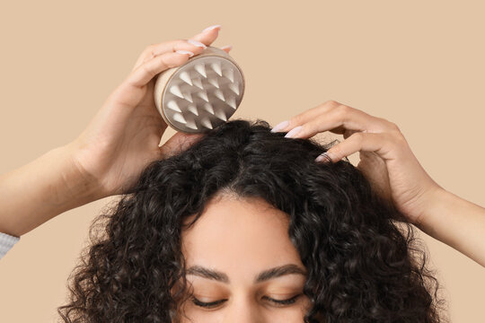 Beautiful young African-American woman using scalp massager for her healthy curly hair on beige background, closeup