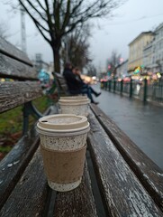 First-person view. A couple is sitting on a bench in a city park, drinking coffee.