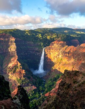A majestic cascading waterfall plunges into a deep canyon, surrounded by lush greenery, rocky peaks, and a blue sky dotted with clouds