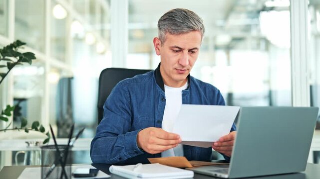 Cheerful male businessman in a blue shirt and white t-shirt opens an important letter in a modern office. Successful male reads the document with happy and excited emotions.