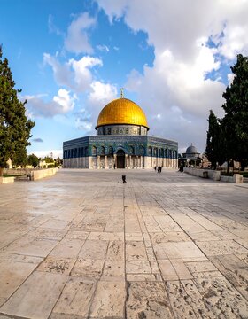 A majestic structure with a golden dome stands under a cloudy sky, surrounded by stone walkways. Trees frame the scene