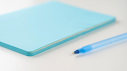 A blue pen rests on top of a blue notebook, both of which are made of paper. The pen is blue and has a small, pointed tip, while the notebook is also blue and has a smooth, glossy surface