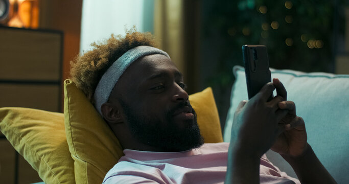 Close-up of young man with curly hair and beard lying on a sofa with yellow pillows. He scrolls on his phone, sees a funny photo of a friend, and laughs out loud. - Powered by Adobe