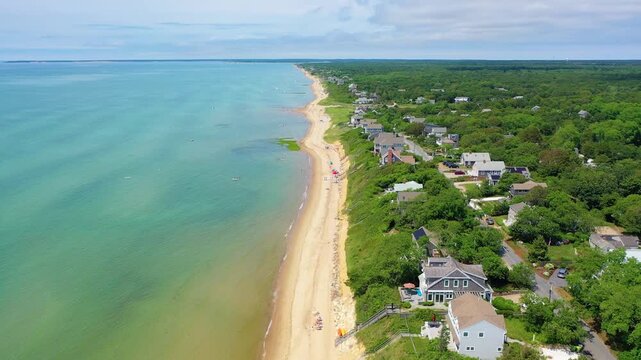 Coastal neighborhood of beach houses nestled in trees overlooks a sandy shoreline where families gather with towels, swimmers enjoy calm surf, and boats scatter across clear blue water.
