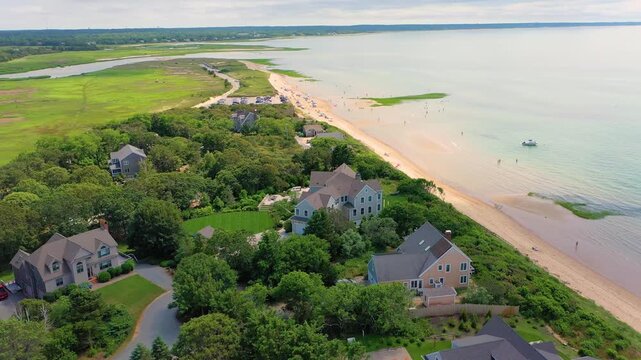 Drone circles a coastal property surrounded by greenery, sandy shoreline filled with families, and turquoise water glowing under the soft radiance of summer.