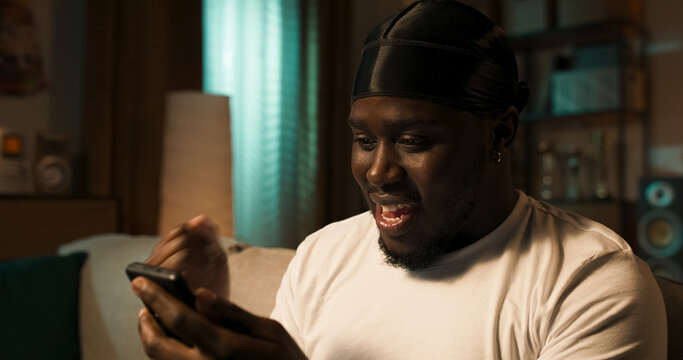 Man lounges on a living room sofa, smiling with excitement. Holding a smartphone, he chats with colleagues and clients online using various social networking platforms.