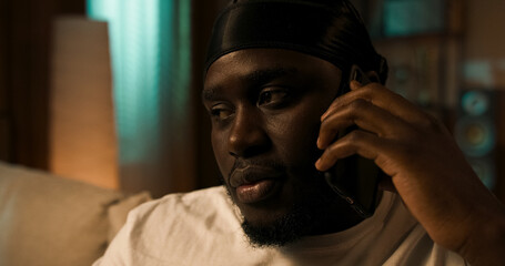 A cheerful young handsome man talking on his cell phone and looking away with a smile, sitting on the couch in the living room.