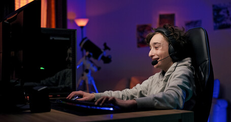 A child gamer focuses on a shooter game with his team, wearing a headset and sitting by the screen in a dark room illuminated by colorful LED lighting.