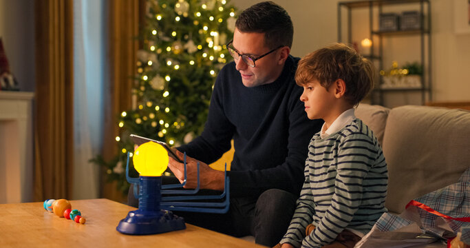 During Christmas evening, a father teaches his child by reviewing toy instructions on a tablet. They arrange planets in proper places on the glowing model of the solar system. - Powered by Adobe