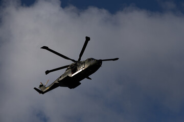 Dark silhouette of a military helicopter flying under a cloudy sky, rotor blades in motion.