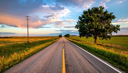 A long straight asphalt road stretches towards the horizon with fields on either side, under a colorful sunset