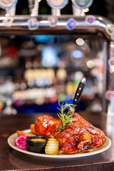 Barbecued meat with sauce, rosemary, and edible flower on ceramic plate. Served with pickles, slaw, and dip. Restaurant setting, dramatic knife presentation.