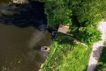 Naklejka premium Aerial view of a wooden dock extending into a dark, murky pond, bordered by dense green trees and grassy banks. A dirt path runs alongside the lush vegetation