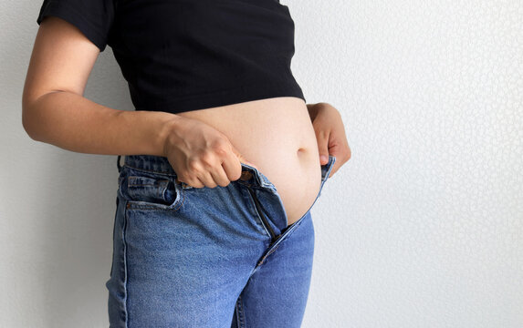 Side view of woman trying to button tight jeans, visible round belly under black shirt, concept of body image and size issues